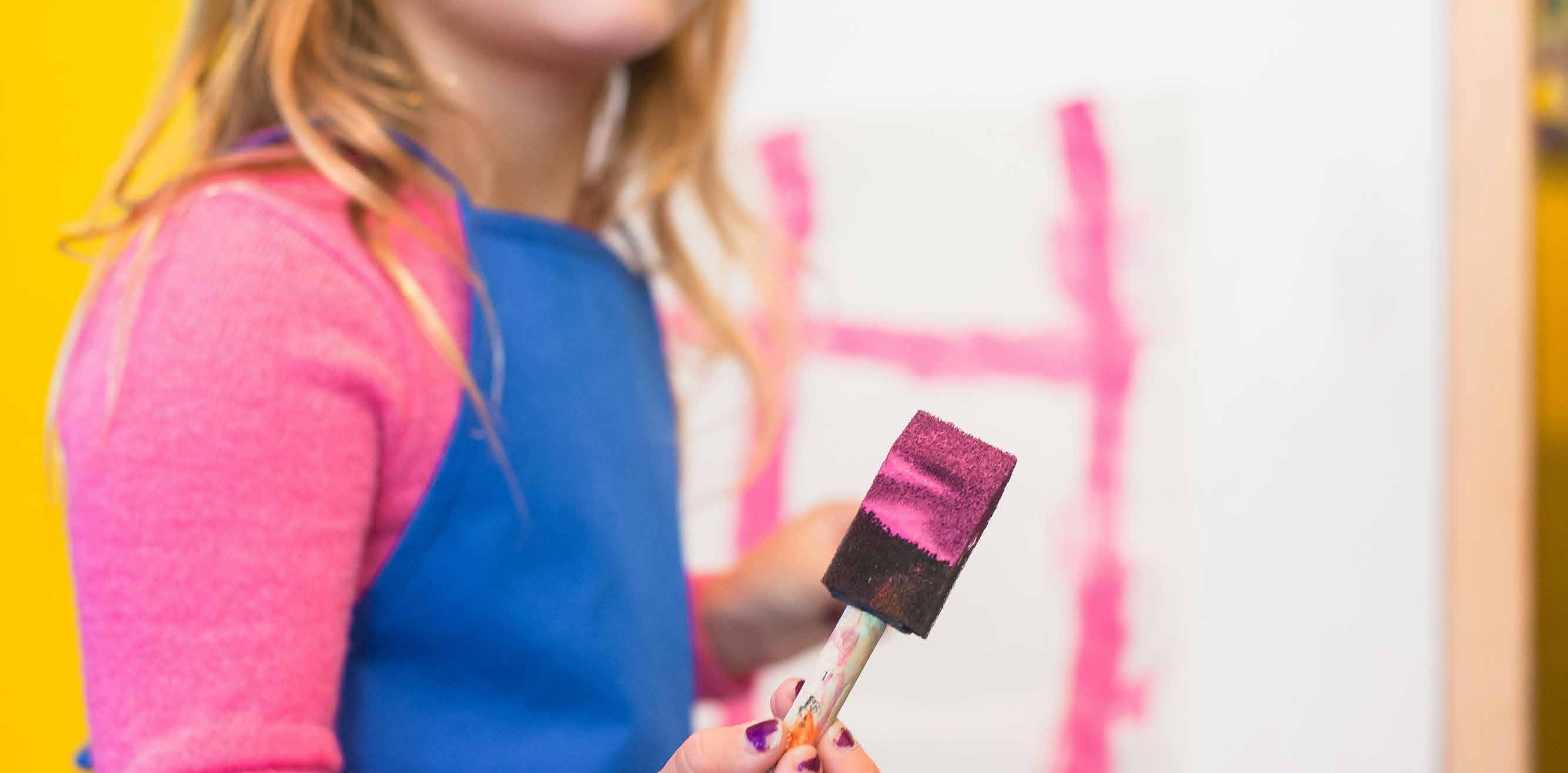 A young child in a blue apron holding a sponge paintbrush covered in pink paint at an early learning centre
